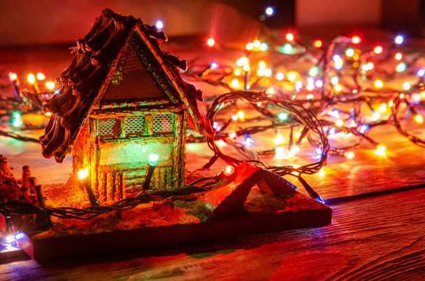 Όμορφο Χριστουγεννιάτικο φόντο. Ξύλινο σπίτι στέκεται σε ξύλινο φόντο ανάμεσα στα φώτα των Χριστουγέννων κοντά - Φωτογραφία, εικόνα