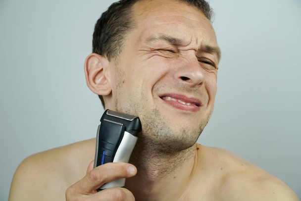 темноволосый красивый мужчина бреется электрической бритвой и делает лица перед камерой. - Фото, изображение