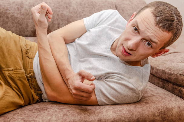 Мужчина лежит на диване в коричневых штанах и белой футболке, у него болит живот.. - Фото, изображение