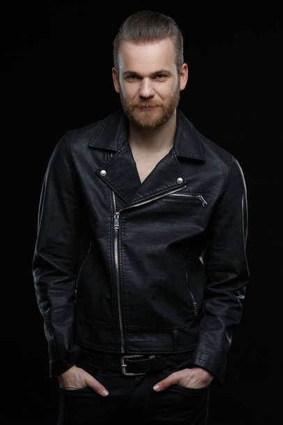 Cute smiling bearded man in a leather jacket isolated on a black background. - Фото, зображення