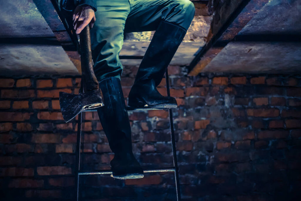 Piedi di un uomo in stivali di gomma con un'ascia tra le mani sulle scale nel seminterrato. il concetto di ladro, assassino, maniaco. vista dal basso - Foto, immagini