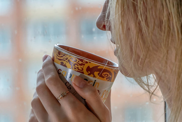 Menina loira desfrutando de seu café da manhã ou chá, olhando através de uma janela chuvosa. Linda menina irreconhecível romântico beber uma bebida quente em uma casa confortável. Humor chuvoso. - Foto, Imagem