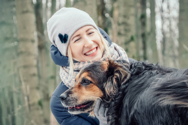 beautiful blonde girl in a gray jacket and a light cap is sitting with a smile near a shepherd dog in the forest. The girl went for a walk in the winter forest with a shepherd dog - Foto, Bild