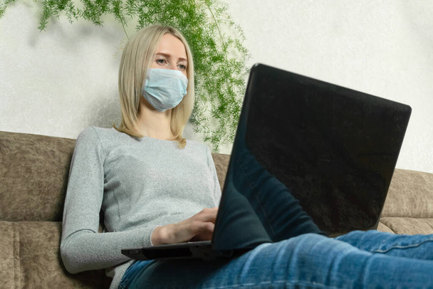 Mujer rubia joven con una máscara médica con un ordenador portátil en su regazo está sentado en el sofá. concepto de autoaislamiento, pandemias. - Foto, imagen