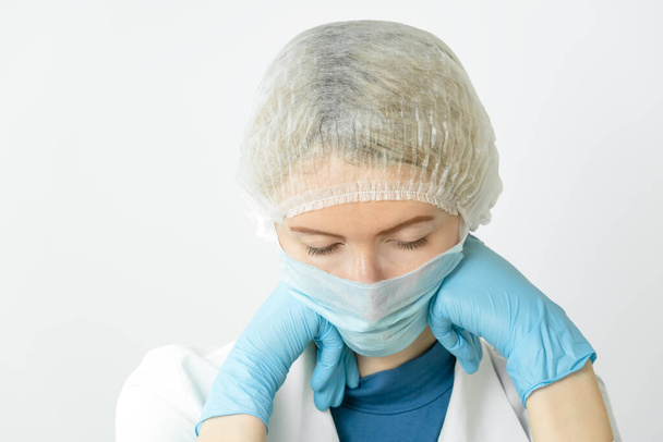 втомлена жінка-лікар у медичній масці та медичній кепці застрягла підборіддям у її блакитних рукавицях та заплющила очі, копіюючи простір
 - Фото, зображення