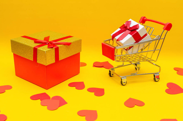 gift box in shopping cart and big gift box on yellow background next to red hearts, copy space. Gift concept, Valentine's Day, holiday - Foto, Imagem