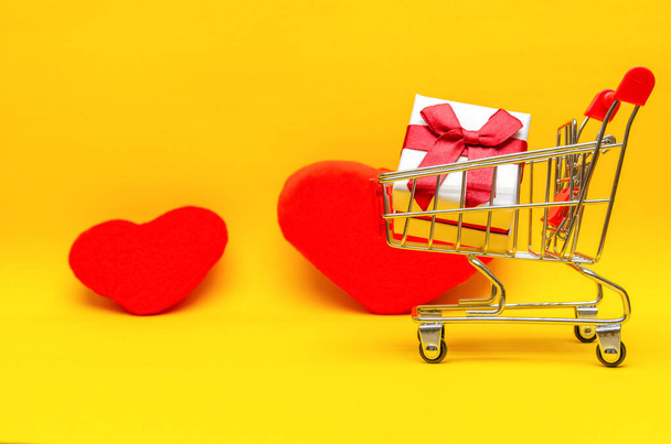 Kırmızı kalplerin yanında sarı arka planda alışveriş arabasında hediye kutusu, kopyalama alanı. Hediye kavramı, Sevgililer Günü, tatil - Fotoğraf, Görsel