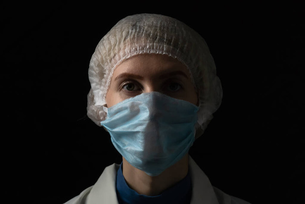 Jonge vrouwelijke arts in medisch masker, medische jurk en pet op zwarte achtergrond met kopieerruimte - Foto, afbeelding