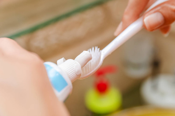 Detailní záběr na dívčí ruce nanášení bílé zubní pasty a bílý kartáček v koupelně nad dřezem - Fotografie, Obrázek