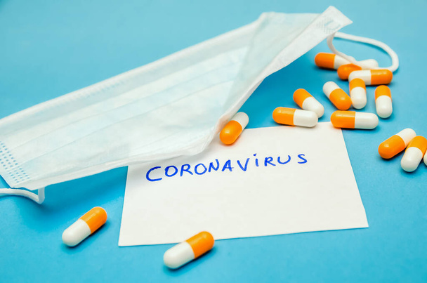Maska medyczna, pomarańczowo-białe kapsułki na niebieskim tle i napis Coronavirus na białym prześcieradle. Pierwsza pomoc, opieka podczas choroby. - Zdjęcie, obraz