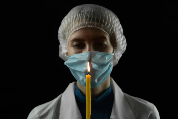 Young woman in a medical mask, medical cap and white coat holds a burning candle in front of her on a black background. Concept of last hope, faith in a bright future. - Photo, Image