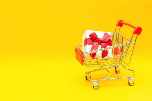 Sarı arka planda alışveriş arabasında hediye kutusu, kopyalama alanı. Hediye kavramı, Sevgililer Günü, tatil - Fotoğraf, Görsel