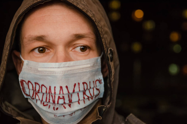 άντρας με κουκούλα με ιατρική μάσκα στο πρόσωπό του με την επιγραφή Coronavirus στο σκοτάδι. Έννοια της υγειονομικής περίθαλψης, ιατρική - Φωτογραφία, εικόνα