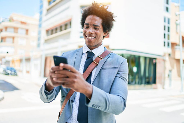 A fiatal afro-amerikai üzletember öltönyt visel, boldogan mosolyog. Álló mosollyal az arcon segítségével smartphone a város utcájában. - Fotó, kép