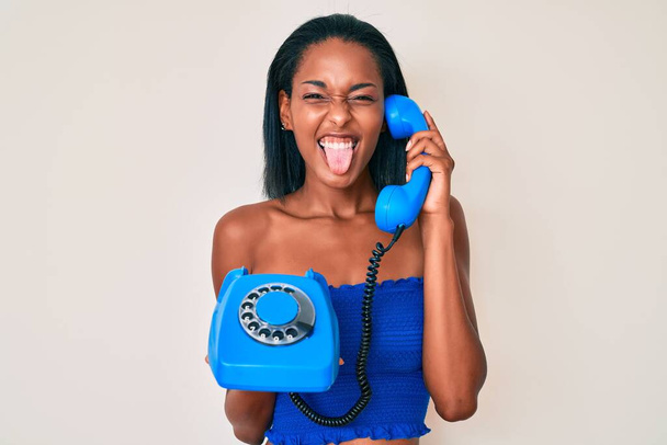 Nuori afrikkalainen amerikkalainen nainen tilalla vuosikerta puhelin törröttää kielen ulos onnellinen hauska ilme.  - Valokuva, kuva