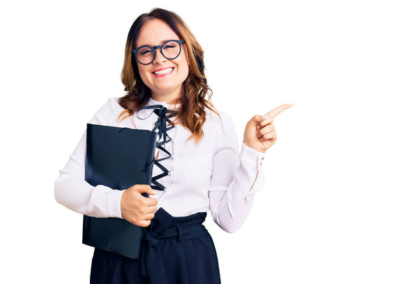 Giovane bella donna caucasica indossa camicia da lavoro e occhiali tenendo cartella sorridente felice indicando con mano e dito di lato  - Foto, immagini