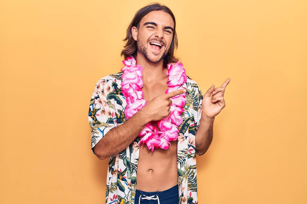 Joven hombre guapo con traje de baño y lei hawaiano sonriendo y mirando a la cámara apuntando con dos manos y dedos hacia un lado.  - Foto, Imagen