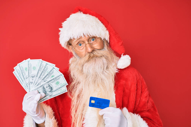 Staruszek w kostiumie Świętego Mikołaja, trzymający dolary i karty kredytowe bez pojęcia i zdezorientowany wyraz twarzy. koncepcja wątpliwości.  - Zdjęcie, obraz