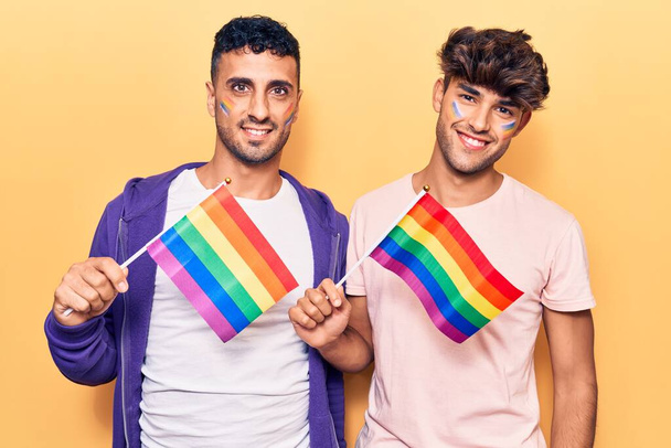 Genç eşcinsel çift ellerinde gökkuşağı lgbtq bayrakları tutarken pozitif ve mutlu görünüyorlar. Dişlerini gösteren bir gülümsemeyle ayakta duruyorlar.  - Fotoğraf, Görsel