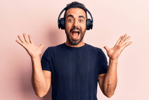 Junger hispanischer Mann hört Musik über Kopfhörer und feiert den Sieg mit glücklichem Lächeln und Siegerausdruck mit erhobenen Händen  - Foto, Bild