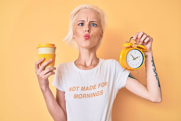 Young blonde woman with tattoo holding alarm clock and take away cup of coffee making fish face with mouth and squinting eyes, crazy and comical.  - Fotoğraf, Görsel