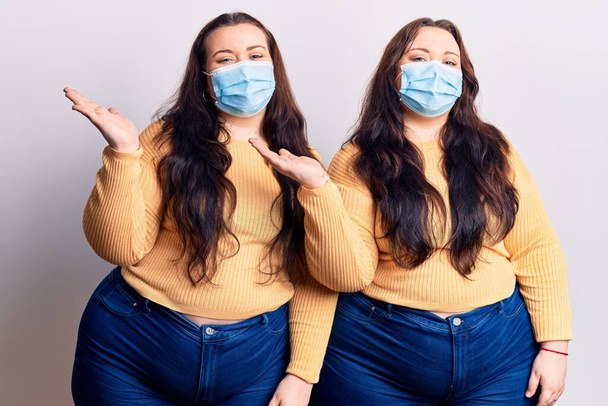 Young plus size twins wearing medical mask smiling cheerful presenting and pointing with palm of hand looking at the camera.  - Photo, Image