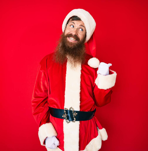 Handsome young red head man with long beard wearing santa claus costume smiling with happy face looking and pointing to the side with thumb up.  - 写真・画像