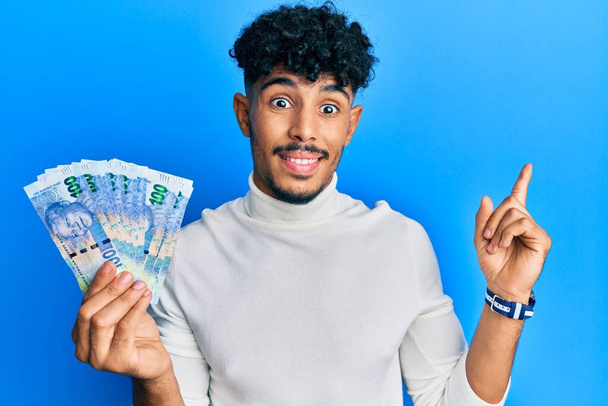 Молодой араб, красивый мужчина с банкнотами 100 рандов, улыбающийся, указывая рукой и пальцем в сторону.  - Фото, изображение