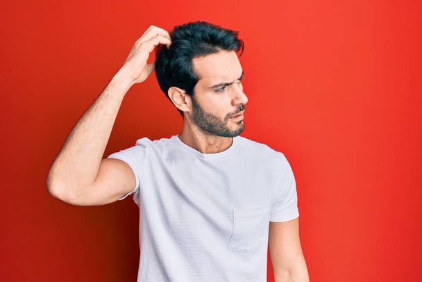 Junger hispanischer Mann in lässigem weißem T-Shirt verwirrt und wundert sich über Fragen. Unsicher im Zweifel, mit der Hand auf dem Kopf denkend. Nachdenkliches Konzept.  - Foto, Bild
