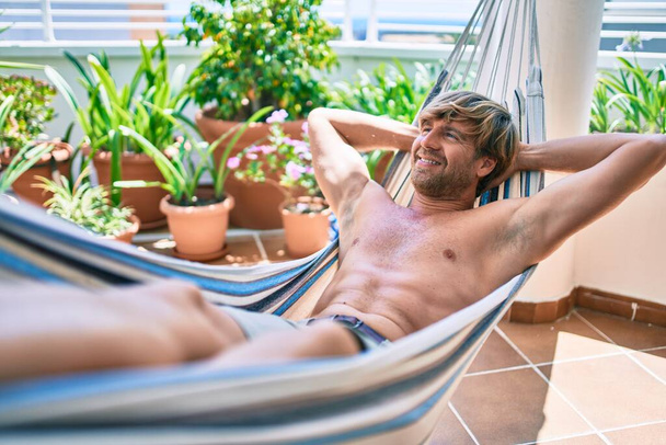 Middelbare leeftijd knappe man op het terras van zijn huis ontspannen liggend shirtloos op een hangmat - Foto, afbeelding