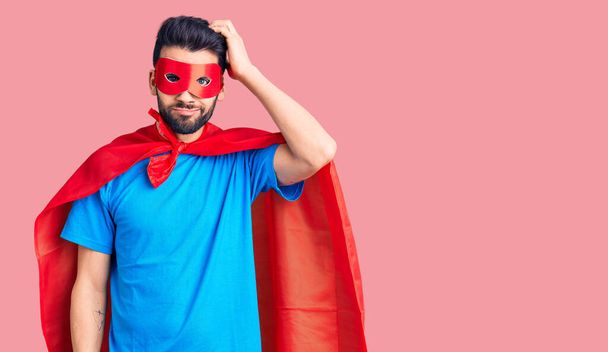 Young handsome man with beard wearing super hero costume confuse and wonder about question. uncertain with doubt, thinking with hand on head. pensive concept.  - Photo, Image