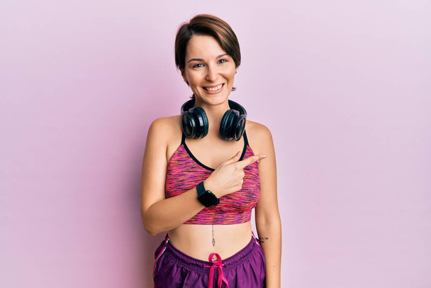Young brunette woman with short hair wearing sportswear and headphones cheerful with a smile of face pointing with hand and finger up to the side with happy and natural expression on face  - Photo, Image
