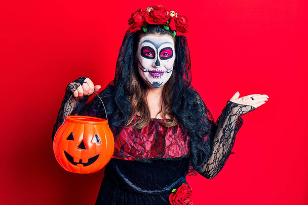 Young woman wearing day of the dead costume holding pumpkin celebrating victory with happy smile and winner expression with raised hands  - Photo, Image