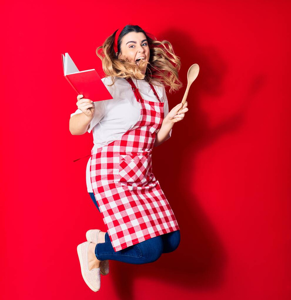 Young beautiful blonde cook woman wearing apron smiling happy. Jumping with smile on face holding recipe book and spoon over isolated red background - Photo, Image