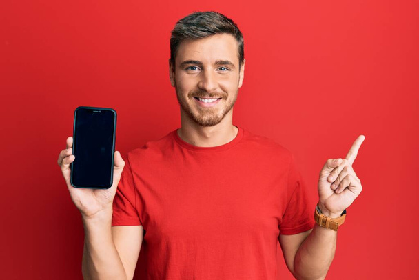 Bonito homem caucasiano segurando smartphone mostrando tela sorrindo feliz apontando com a mão e o dedo para o lado  - Foto, Imagem