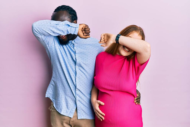 Young interracial couple expecting a baby, touching pregnant belly smiling cheerful playing peek a boo with hands showing face. surprised and exited  - Fotografie, Obrázek