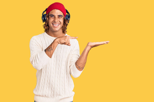 Jeune homme hispanique écoutant de la musique à l'aide d'écouteurs étonnés et souriants à la caméra tout en présentant avec la main et pointant du doigt.  - Photo, image