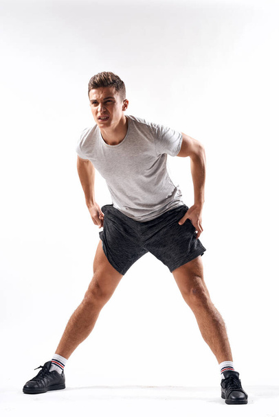 A sports man in shorts and a full-length T-shirt does exercises on a light background - Fotoğraf, Görsel