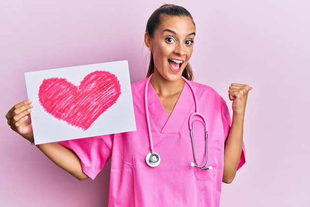 Jeune femme hispanique portant l'uniforme de médecin tenant le coeur tirant pouce pointant vers le côté souriant heureux avec la bouche ouverte  - Photo, image