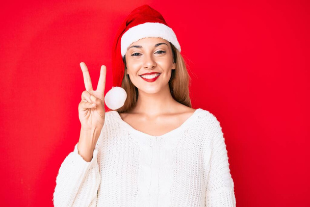 A fiatal barna nő karácsonyi kalapban mosolyog, boldog arccal kacsint a kamerára, győzelmi jelet adva. Második..  - Fotó, kép