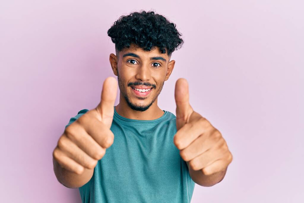 Junger arabischer gutaussehender Mann in lässiger Kleidung, der positive Gesten mit der Hand zustimmt, lächelnd die Daumen nach oben streckt und sich über den Erfolg freut. Siegergeste.  - Foto, Bild