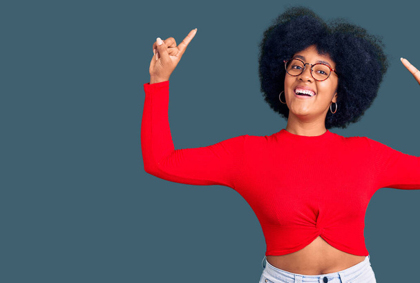 Fiatal afro-amerikai lány alkalmi ruhát és szemüveget visel mosolyogva csodálkozva és meglepve, és rámutatva ujjakkal és felemelt karokkal.  - Fotó, kép