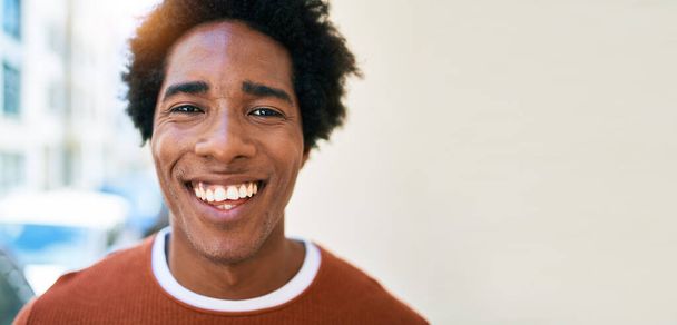 Fiatal, jóképű afro-amerikai férfi, alkalmi ruhákat visel, boldogan mosolyog. Mosollyal az arcán állni a város utcáján.. - Fotó, kép