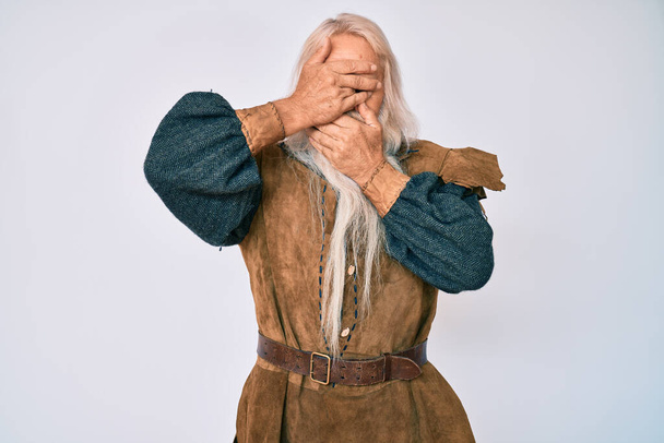 Γέρος ηλικιωμένος άνδρας με γκρίζα μαλλιά και μακριά γενειάδα φορώντας viking παραδοσιακή φορεσιά που καλύπτει τα μάτια και το στόμα με τα χέρια, έκπληκτος και σοκαρισμένος. απόκρυψη συγκίνησης  - Φωτογραφία, εικόνα