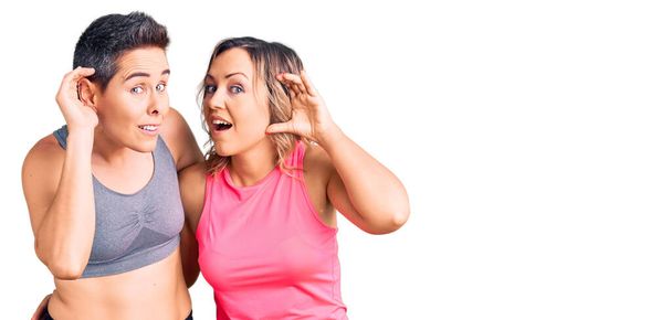 Een paar vrouwen met sportkleding glimlachend met hand op oor luisterend naar geruchten of roddels. doofheid.  - Foto, afbeelding