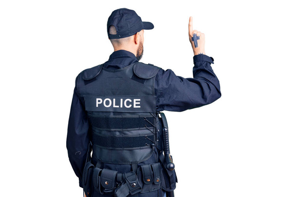 Joven hombre guapo con uniforme policial posando hacia atrás apuntando hacia adelante con la mano del dedo  - Foto, imagen