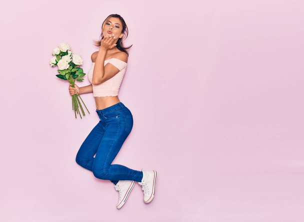Young beautiful girl holding bouquet of flowers. Throwing kiss with hand jumping over isolated pink background - Foto, Bild
