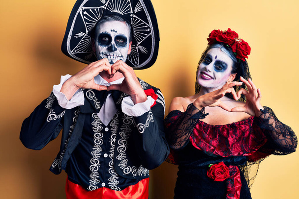 Młoda para ubrana w meksykański dzień martwego kostiumu na żółtym uśmiechu zakochana robiąca ręcznie kształt symbolu serca. romantyczna koncepcja.  - Zdjęcie, obraz