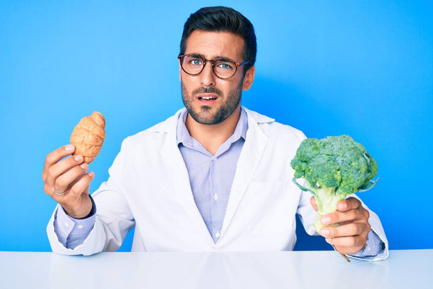 Young hispanic man as nutritionist doctor holding croissant and broccoli clueless and confused expression. doubt concept.  - Photo, Image