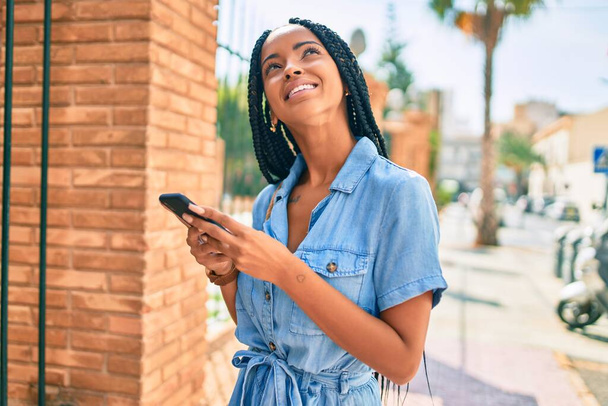 Joven mujer afroamericana sonriendo feliz usando smartphone en la ciudad. - Foto, imagen